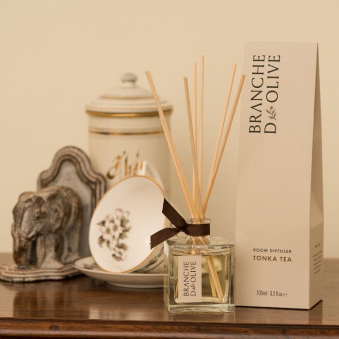 Branche d'Olive Room Diffuser in Tonka Tea fragrance on a wooden table with tea cup and saucer, tea caddy and decorative wooden ornament