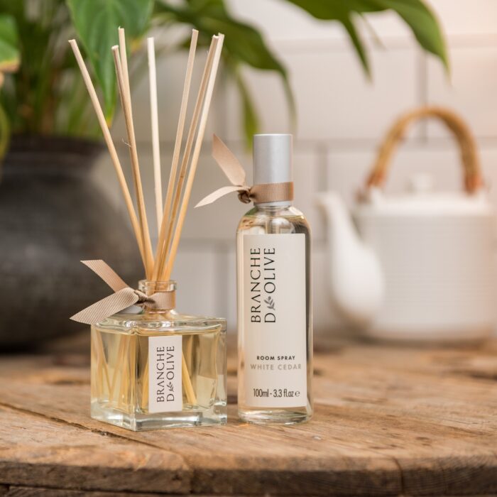 A Branche d'Olive unboxed diffuser and room spray in White Cedar fragrance shown on a wooden table with a white china teapot in the distance.in