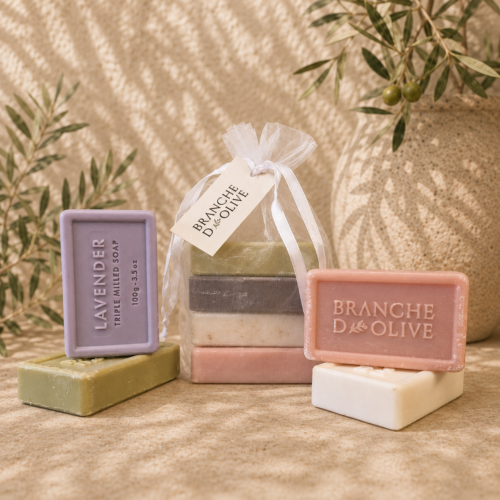 Branche d'Olive guest soaps. Four are shown bagged in white voile with a hanging tag and four are shown outside the bag. The image is shown in bright sunlight set against a backdrop of olive branches.