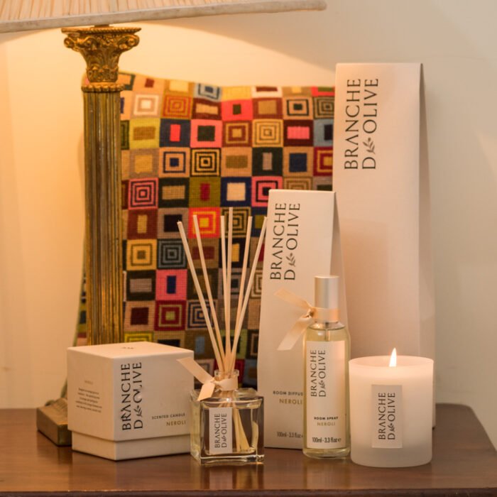 Branche d'Olive Neroli scented Room Diffuser, Room Spray and Candle on a wooden table in front of a tapestry cushion and beside a brass lamp
