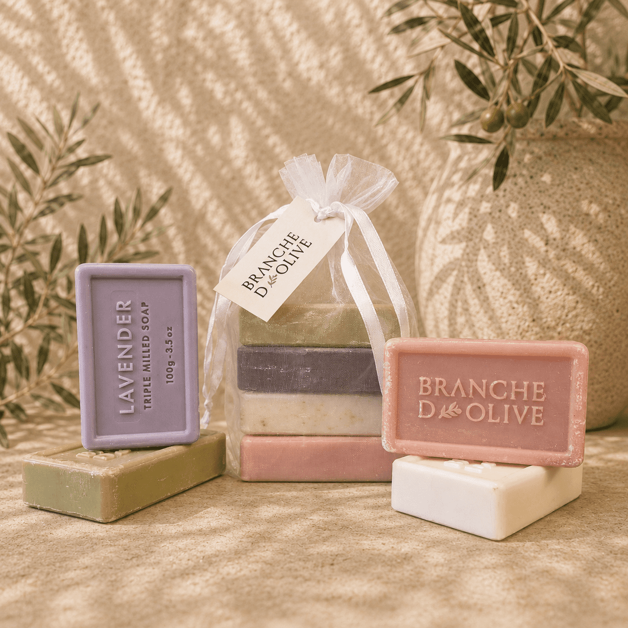Branche d'Olive guest soaps. Four are shown bagged in white voile with a hanging tag and four are shown outside the bag. The image is shown in bright sunlight set against a backdrop of olive branches.