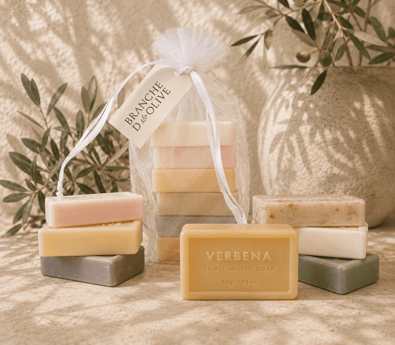 Branche d'Olive guest soaps. Six are shown bagged in white voile with a hanging tag and a group are shown outside the bag. The image is shown in bright sunlight set against a cream wall with olive branches in a pot and freestanding.